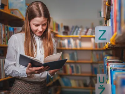 Schoolgirl searching and choosing books from a bookshelf in the library. Education, literature, and new edition concepts.