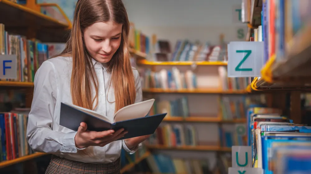 Schoolgirl searching and choosing books from a bookshelf in the library. Education, literature, and new edition concepts.