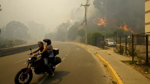 Residents evacuate on a motorcycle as smoke caused by forest fires fill the sky and flames spread into Vina del Mar, Chile, Saturday, Feb. 3, 2024. Officials say intense forest fires burning around a densely populated area of central Chile have left several people dead and destroyed hundreds of homes. (AP Photo/Esteban Felix)