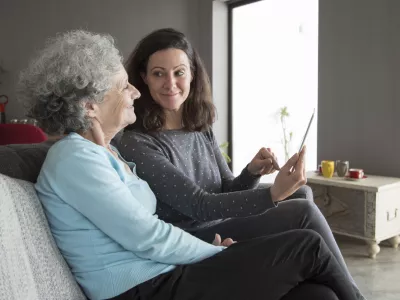 Smiling senior woman and her daughter using tablet computer. Mother and daughter sitting on couch with home interior in background. Technology concept. / Foto: Mangostar_studio