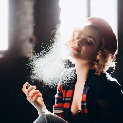 A beautiful blonde girl in a red beret holds a small bottle in her hands spraying perfume on herself enjoys armature. Backlight, soft focus.