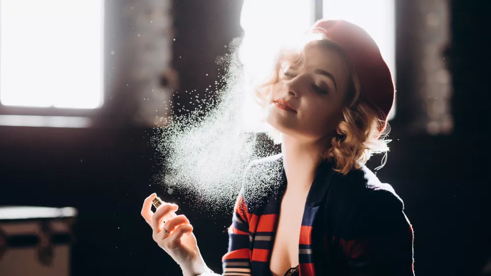 A beautiful blonde girl in a red beret holds a small bottle in her hands spraying perfume on herself enjoys armature. Backlight, soft focus.