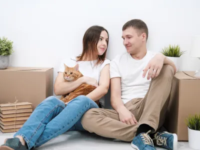 A young married couple with a red cat, a man and a woman, are sitting on the floor in a bright room against the background of cardboard boxes, green plants and things. Concept themes moving to a new home.