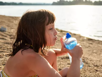 A girl drinks water while sitting on the beach by the lake during a summer day. / Foto: Marizza