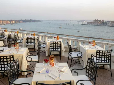 The rooftop terrace of Restaurant Terrazza Danieli at sunset. Small, square, candlelit tables dressed in tablecloths epitomise fine dining in Venice. Spectacular views overlook the Venetian lagoon with tradtional gondolas. 