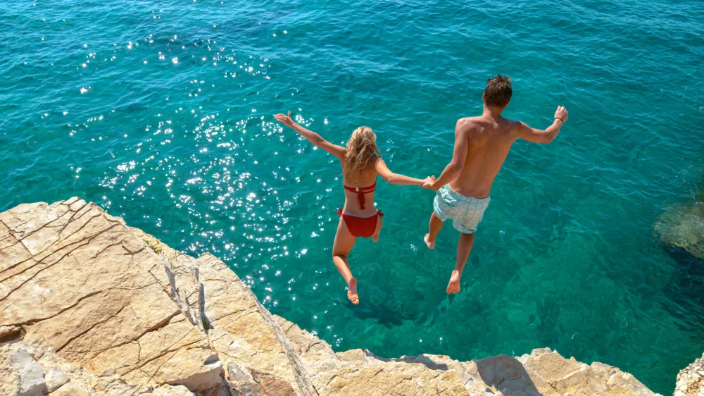 Carefree tourists hold hands while jumping into the refreshing blue sea during a relaxing summer vacation. Active young woman and her boyfriend dive off a high cliff and into the deep blue ocean.