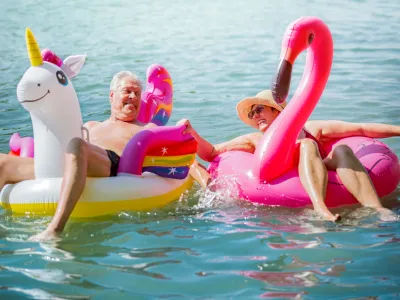 Elderly couple having fun on inflatable flamingo and unicorn. Funny active pensioners happy together enjoying summer vacation on the beach in Europe, laughing, playing the fool, splashing water.