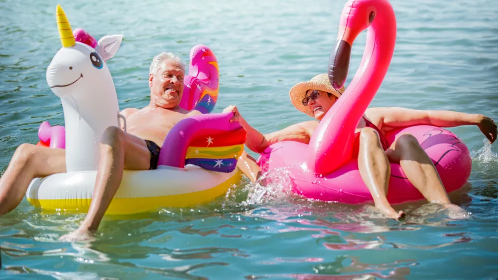 Elderly couple having fun on inflatable flamingo and unicorn. Funny active pensioners happy together enjoying summer vacation on the beach in Europe, laughing, playing the fool, splashing water.