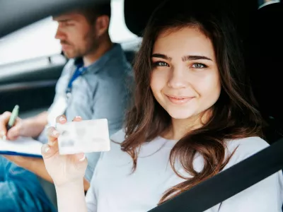 2D815X2 Attractive young woman showing driver's licence, looking at camera and smiling. Brunette woman sitting on drivers seat with fastened seat belt. Blurre