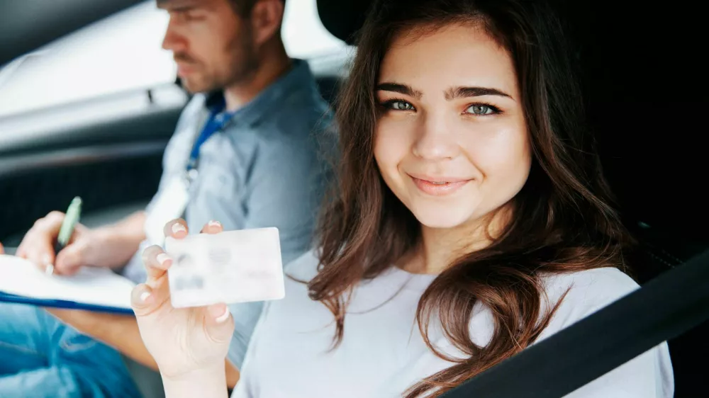 2D815X2 Attractive young woman showing driver's licence, looking at camera and smiling. Brunette woman sitting on drivers seat with fastened seat belt. Blurre