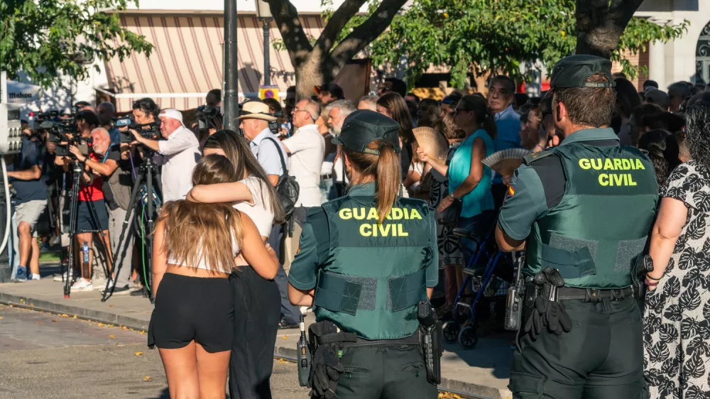 18 August 2024, Spain, Mocejon: Two Guardia Civil officers watch two girls embrace during the minute of silence for the murder of a minor at the door of the Mocejon Town Hall. The Mocejon City Council has ordered three days of national mourning for the murder of an 11-year-old minor who was playing soccer this morning with other children on the municipal soccer pitch "Angel Tardio" when he was attacked with a sharp object. Photo: Juan Moreno/EUROPA PRESS/dpa