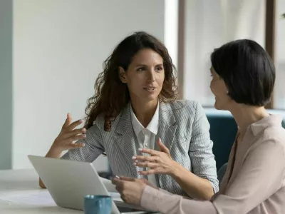 Two business women sit at desk discuss project details, diverse female colleagues met in office, share opinion, working on collaborative task, sales manager makes commercial offer to company client / Foto: Fizkes