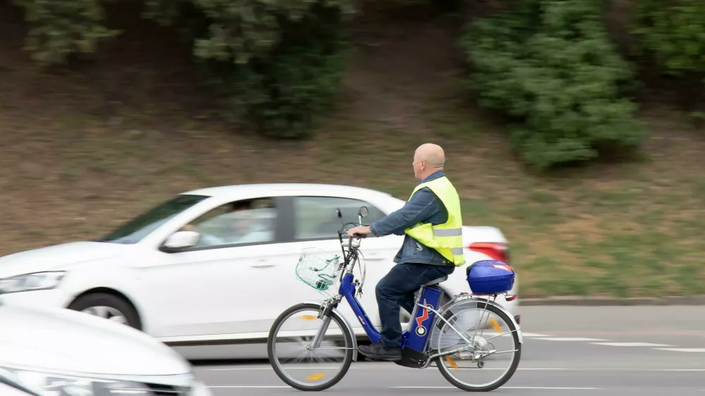 2FP3753 Belgrade, Serbia - May 14, 2021: One senior man wearing fluorescent vest riding an electric bike in city street traffic by the park