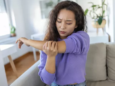 unhappy asian woman with elbow pain indoors. office syndrome health care concept. upset frowning asian lady confused looking at arms hurting sitting on couch at home in cozy bright apartment. / Foto: Stefanamer