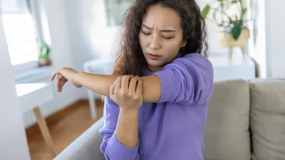 unhappy asian woman with elbow pain indoors. office syndrome health care concept. upset frowning asian lady confused looking at arms hurting sitting on couch at home in cozy bright apartment. / Foto: Stefanamer