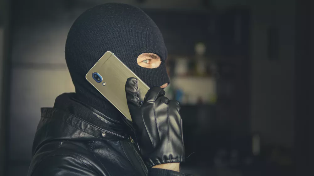 Young adult in black clothes with hidden face. Ill-intended fraudster uses mobile. Fraudster calls. Mobile racket. Hacker hijacks by phone. Cellphone account fraud. Scam / Foto: Diy13