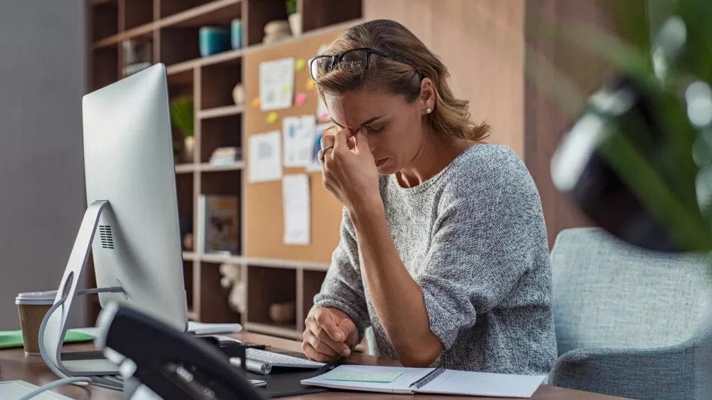 ﻿Exhausted businesswoman having a headache in modern office. Mature creative woman working at office desk with spectacles on head feeling tired. Stressed casual business woman feeling eye pain while overworking on desktop computer.