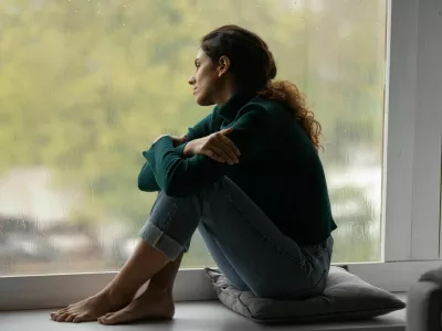 Pensive sad millennial Hispanic woman sit on windowsill at home look in window distance thinking dreaming. Thoughtful young Latino female loner or outcast feel lonely relax rest indoors on rainy day. / Foto: Fizkes