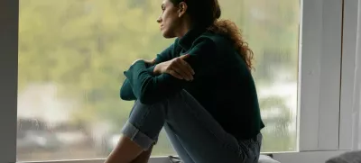 Pensive sad millennial Hispanic woman sit on windowsill at home look in window distance thinking dreaming. Thoughtful young Latino female loner or outcast feel lonely relax rest indoors on rainy day. / Foto: Fizkes