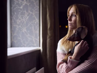 Woman With Pet Dog Trying To Keep Warm By Radiator At Home During Cost Of Living Energy Crisis