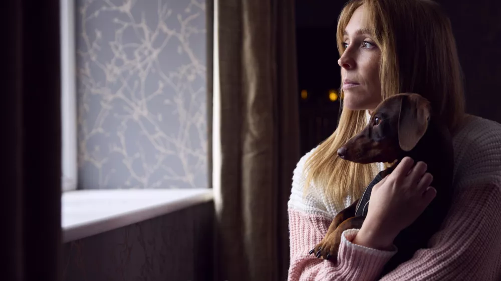 Woman With Pet Dog Trying To Keep Warm By Radiator At Home During Cost Of Living Energy Crisis