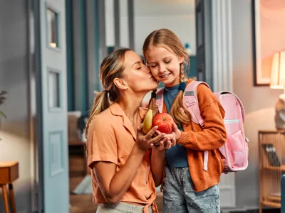 Healthy snack for pupil. Adult woman giving pretty daughter with pink schoolbag apple and banana and kissing her in cheek. Casual female taking care about nutritious meal between lesson for child. / Foto: Harbucks