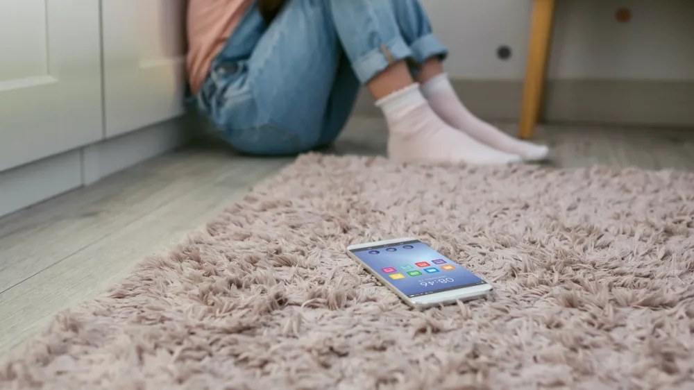 Mobile phone lying on the floor with unrecognizable sad little girl sitting behind. Selective focus on mobile in foreground / Foto: Doble-d