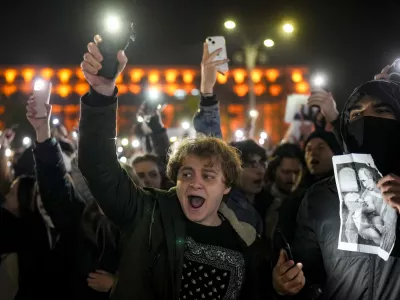 Youngsters shout slogans and flash the light of their mobile phones in Bucharest, Romania, Wednesday, Nov. 27, 2024, next to an altered version of a classic painting, depicting Russian President Vladimir Putin and Calin Georgescu, the independent candidate for Romanian presidency who won the first round of elections making it to the Dec. 8, runoff. (AP Photo/Vadim Ghirda)