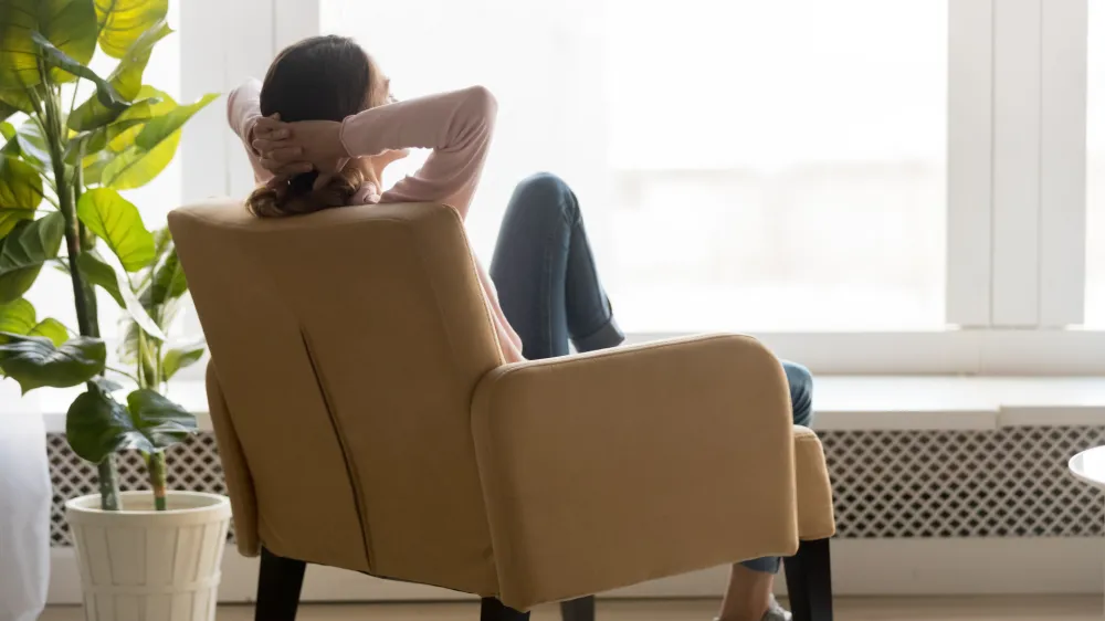 Back view of young happy woman sitting in comfortable armchair, facing shiny window, crossing hands behind head, having rest after housework, resting on weekend, lazy day concept.