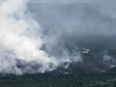 A helicopter carrying a water basket flies past a smoke plume near Lebel-sur-Quevillon, Quebec, Wednesday, July 5, 2023. (Adrian Wyld/The Canadian Press via AP)