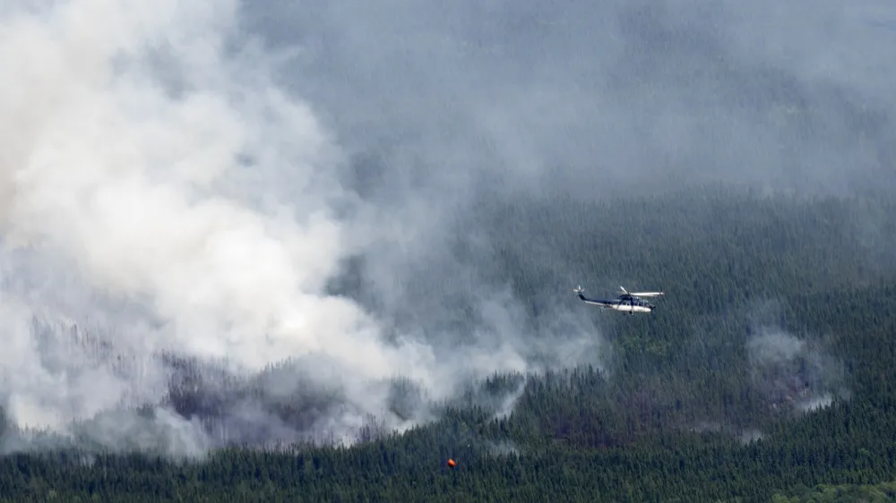 A helicopter carrying a water basket flies past a smoke plume near Lebel-sur-Quevillon, Quebec, Wednesday, July 5, 2023. (Adrian Wyld/The Canadian Press via AP)