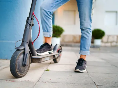 ﻿Close up on woman legs feet standing on the electric kick scooter on the pavement wearing jeans and sneakers in summer day