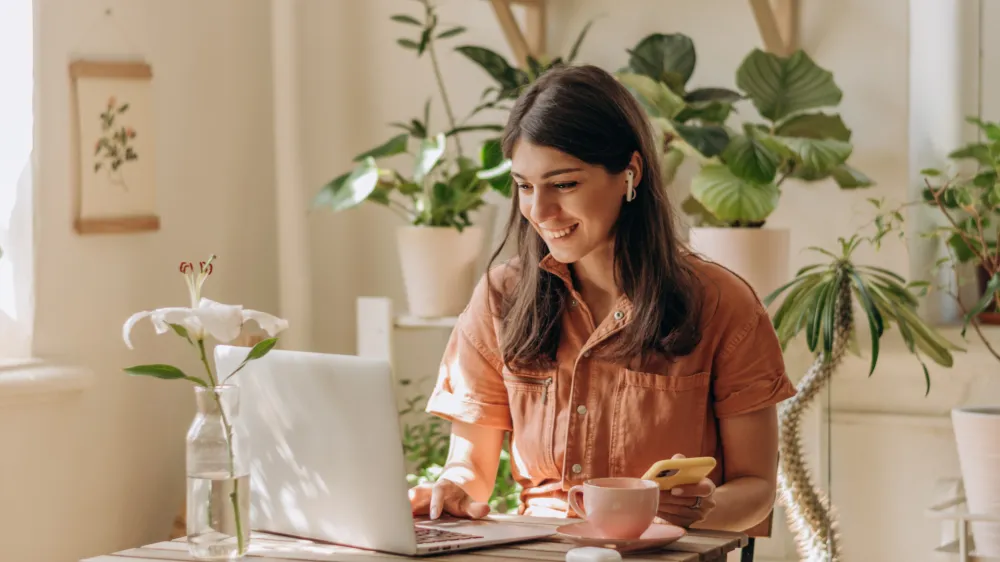 Positive young mixed race woman using a laptop and smartphone at home.Cozy home interior with indoor plants.Remote work, business,freelance,online shopping,e-learning,urban jungle concept.