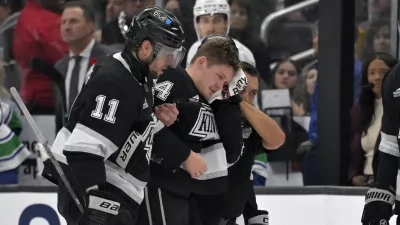 Los Angeles Kings defenseman Mikey Anderson, center, is helped off the ice by Los Angeles Kings center Anze Kopitar (11) after he was hit in the head with the puck during the second period of an NHL hockey game against the Vancouver Canucks, Thursday, Nov. 7, 2024, in Los Angeles. (AP Photo/Jayne-Kamin-Oncea)